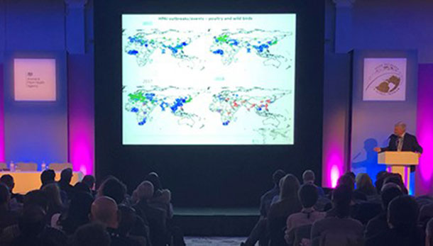 Ian Brown standing at a lectern at the front of an audience with four world maps projected on a large screen.