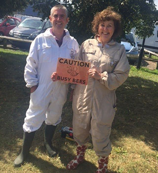 Mark and Sue in their bee-keeping suits holding a sign saying, 'caution, busy bees'.