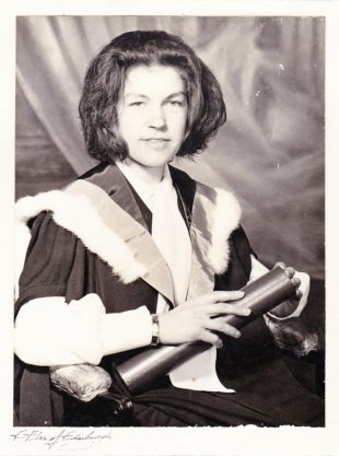 Picture of Jane Clarke at her graduation in black and white. She is wearing a gown and holding a tube,