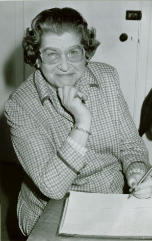 Picture of Lorna Timms in black and white. She is looking at the camera while resting her chin on one hand and holding a pen to paper with the other hand.
