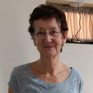 Image of a lady in a light blue top and glasses smiling and looking at the camera with a window and drawn curtains in the background.