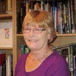 Image of a lady in a purple top and glasses, smiling at the camera in front of a bookshelf full of books.