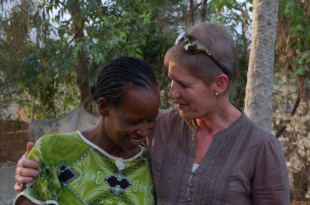 Image of a lady with her arm around another lady with trees in the background, both smiling.