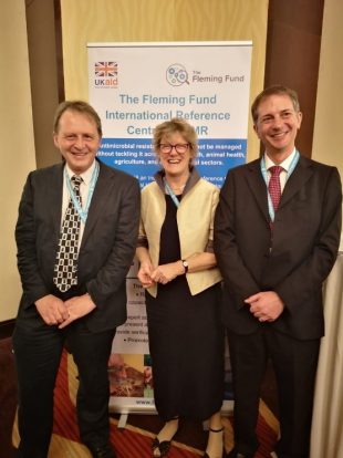 Two gentlemen standing either side of a lady in front of a sign saying, The Fleming Fund, International Reference Centre for AMR