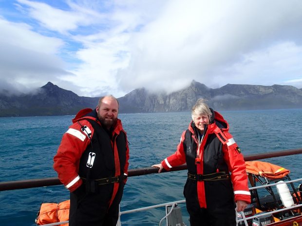 Picture of Jill and a South Georgia biosecurity officer on a boat with mountains and clouds behind them.