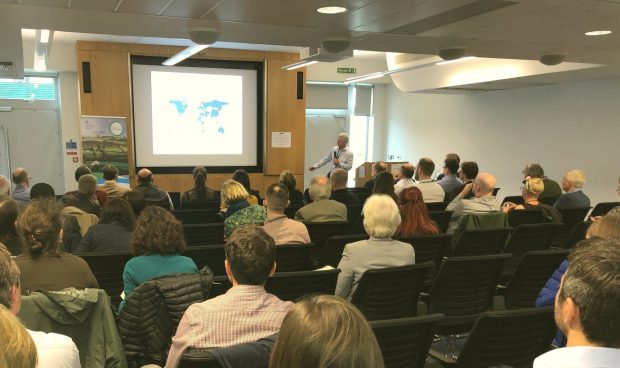 Room of people sitting with their backs to the camera towards a screen depicting a map of the world. Male presenter is standing at the front of the audience.