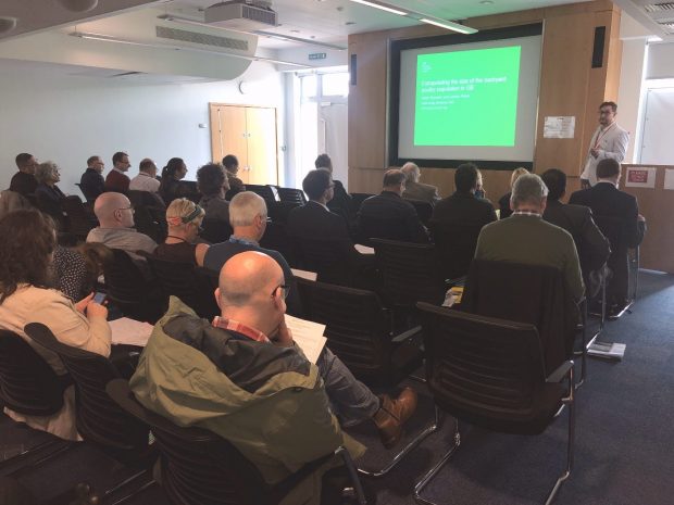 Room of people facing towards a screen where a man is presenting to them.