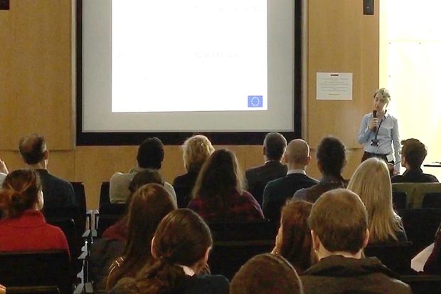 Female standing beside a presentation screen at the front of an audience, giving a presentation