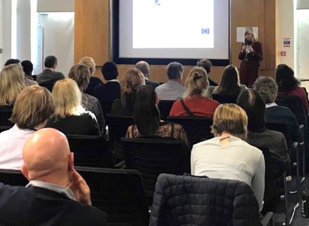 Female standing beside a presentation screen at the front of an audience, giving a presentation