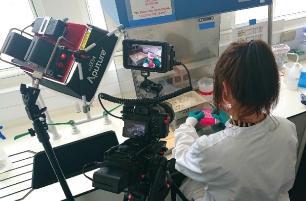 Female scientist performing an experiment while a camera films her from over her shoulder.