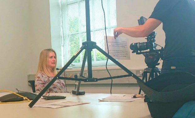 Female sitting behind a desk reading a script held by a male who is also filming her.