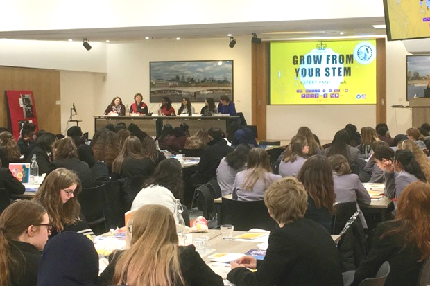 Image of 6 females sitting at a desk on a stage with tables filled with school-aged girls in the audience. A presentation screen reads ' Grow from your STEM'