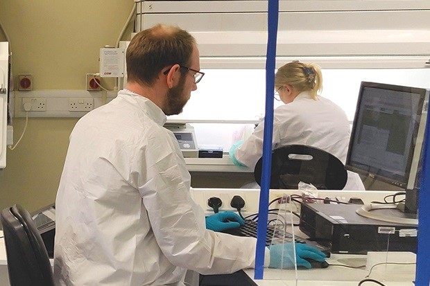 Image of a gentleman scientist in a white coat and gloves working on a computer with a female scientist in the background in a white coat with her back to the camera