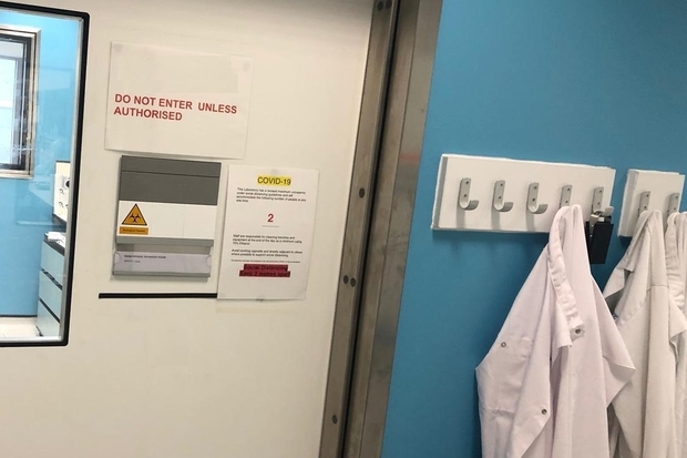 Image of the exterior of a laboratory door with white lab coats hanging up outside. The lab door has signs saying: DO not enter unless authorised and COVID-19.