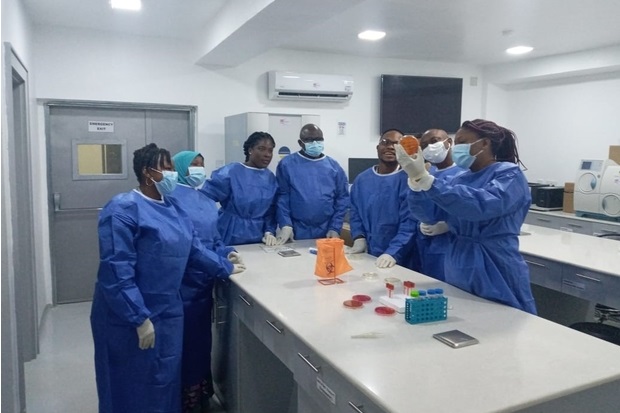 Image of 7 individuals wearing blue overalls and masks in a laboratory looking at another individual holding up a petri dish