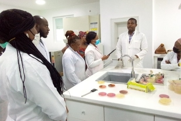 Image of individuals in white coats listening to a male in a white lab coat talking to them.