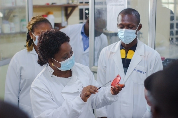 Image of a female wearing a white lab coat holding a petri dish and swab demonstrating to other individuals wearing white coats around her