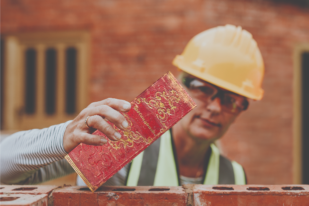 Image of a female bricklayer building a brick wall with a book in her laying hand.