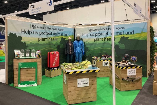 Image of the APHA stand with the backdrop saying, "Help us protect animal and plant health" with four plinths, two mannequins and a red suitcase.