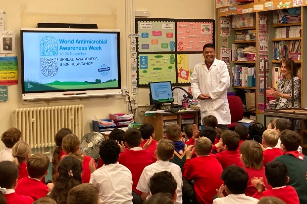 Image of a male in a white lab coat standing in a classroom of children who are sitting on the floor in front of him. There is a screen at the front which says, "World Antimicrobial Awareness Weekn 18-24 November, Spread Awareness Stop Resistance"