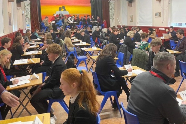 Image of school pupils at small desks across from adults in a school hall