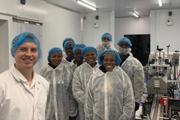 Nine individuals in a group photo wearing white cover-alls and blue hair nets. Some machinery is also visible.