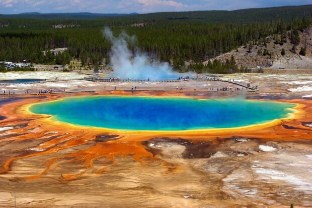 Image of a blue pool of water with colourful hues of yellow, and orange spanning around its circumference. There are people stood next to this feature with trees in the background.