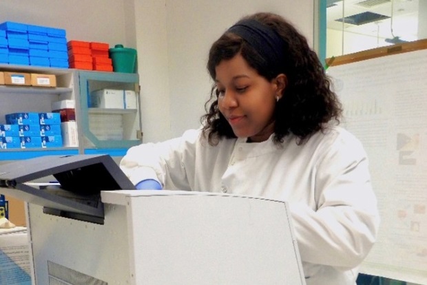 Female scientist in a white laboratory coat and blue gloves operating a small white machine in a laboratory.