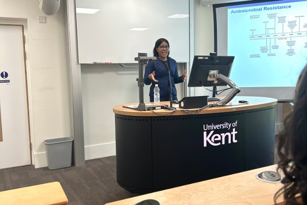 Female standing at a lectern with a computer monitor speaking to an audience in a lecture theatre.