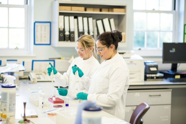 Two female scientists in white laboratory coats, green gloved and safety goggles holding pipettes in a laboratory setting.