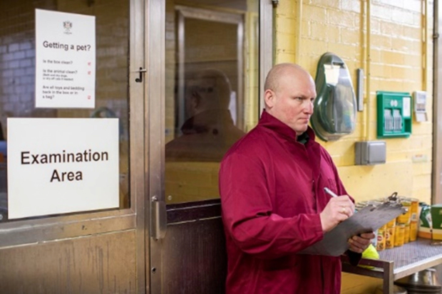 Image of a male dressed in a red boiler suit writing on a clipboard in a room with, "examination area" written on a sign on the door