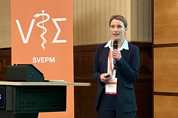 Female speaking into a microphone, next to a lectern and poster which says, "SVEPM"