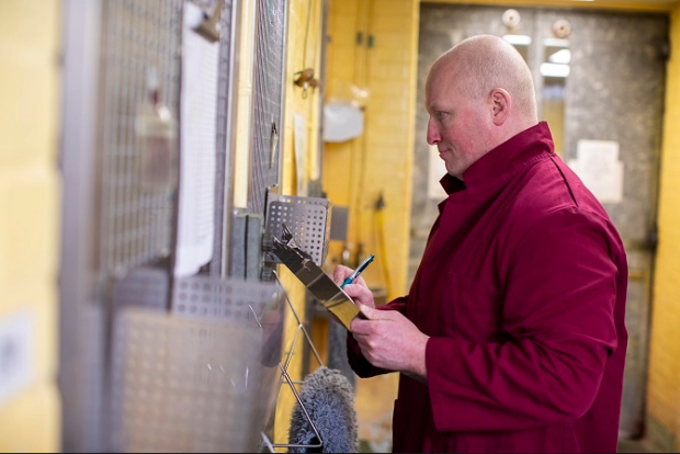 Gentleman in a red jacket, holding a clipboard looking through a grate