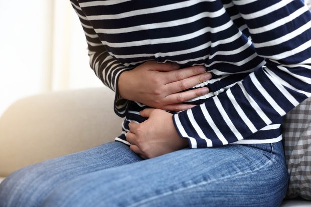 Person in a navy-and-white striped shirt and jeans clutching their abdomen while seated, conveying stomach pain, discomfort, or cramps at home.
