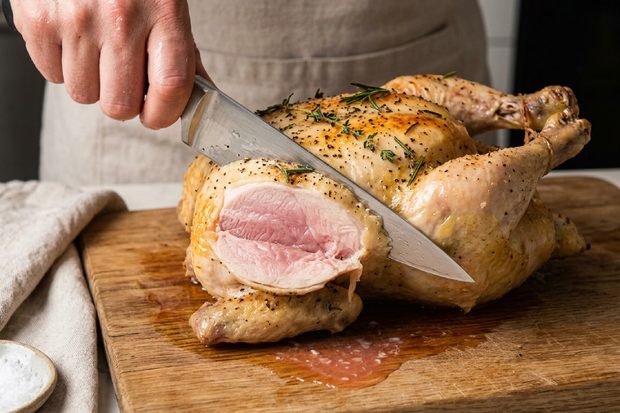 Close-up of a roasted chicken being carved on a wooden board, revealing pink meat, herbs, golden skin, and savoury cooking juices.