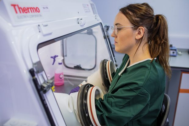 A scientist in teal scrubs and glasses sits in profile by a modern laboratory imaging machine, focused and calm; Thermo branding appears on the equipment.