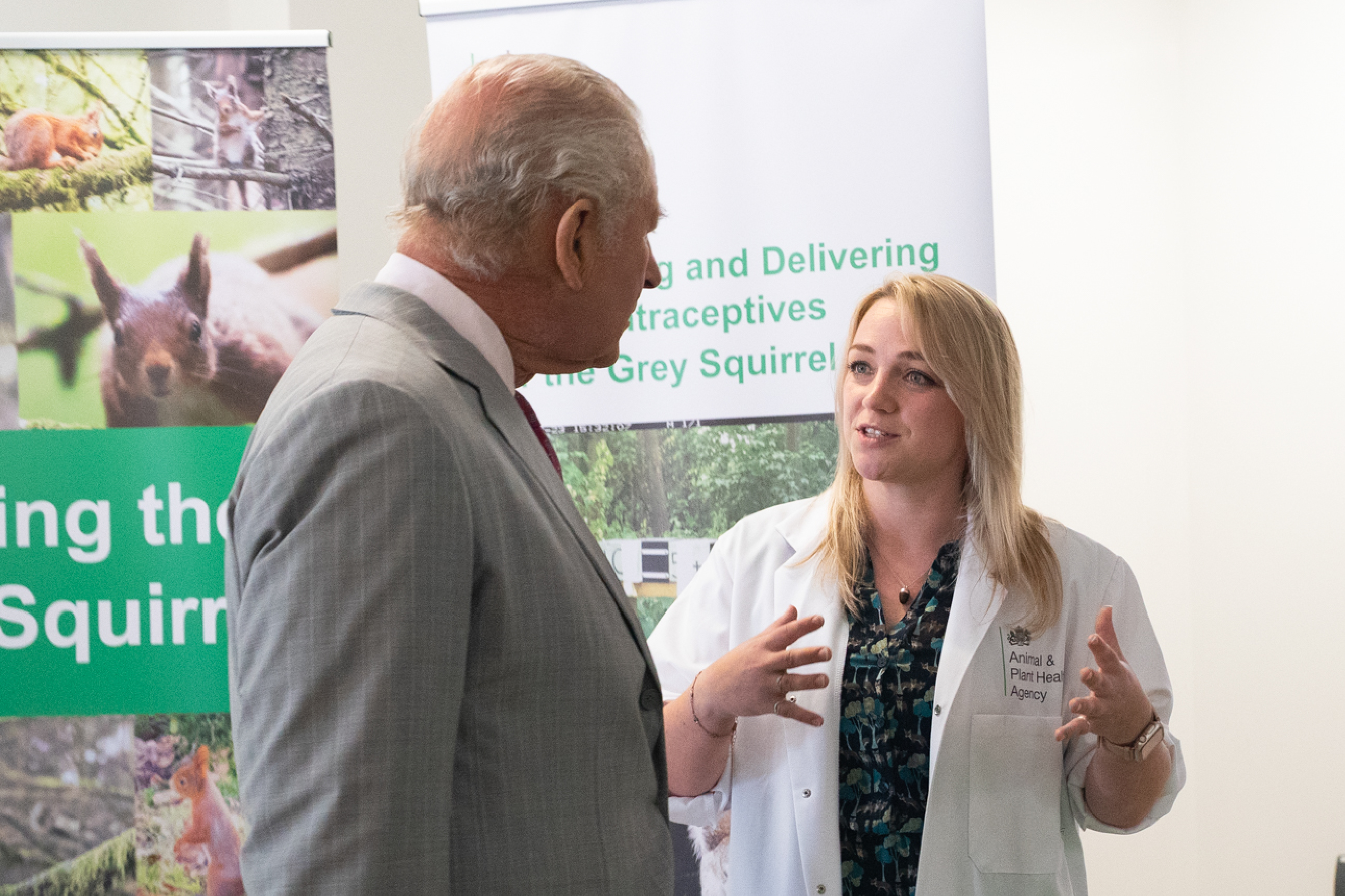 His Majesty The King speaks with a female scientist in a lab coat beside posters about grey squirrel contraceptives; their conversation feels engaged.