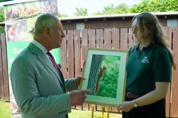 His Majesty The King in a grey suit talks with an APHA staff member holding a framed red squirrel photo; their calm exchange takes place outdoors.
