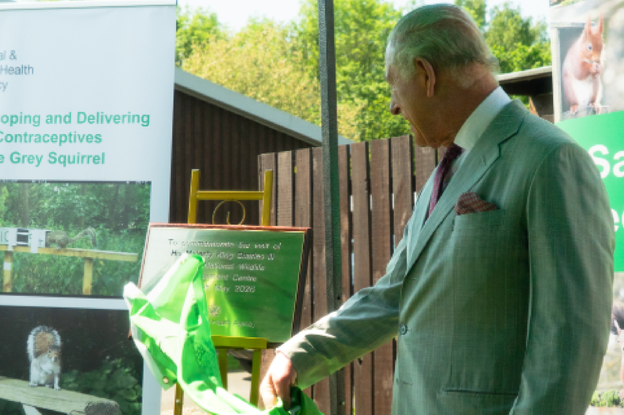 His Majesty The King, wearing a pale grey suit reaches toward a bright green display at an outdoor wildlife exhibit, beside signs about grey squirrels.